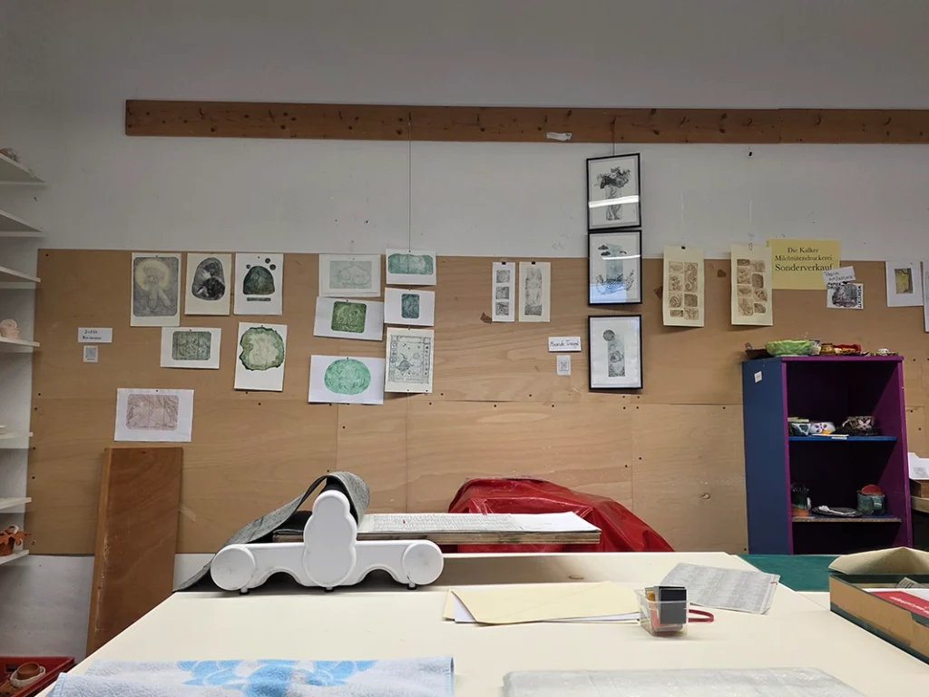 Blick in eine helle Druckwerkstatt mit einer weißen Druckerpresse auf einem Tisch im Vordergrund und zahlreichen Kunstwerken, die an einer Holzwand im Hintergrund hängen.