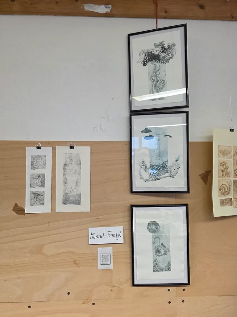 Nahaufnahme einer hölzernen Ausstellungswand. Zu sehen sind mehrere gerahmte und ungerahmte TetraPak-Drucke.